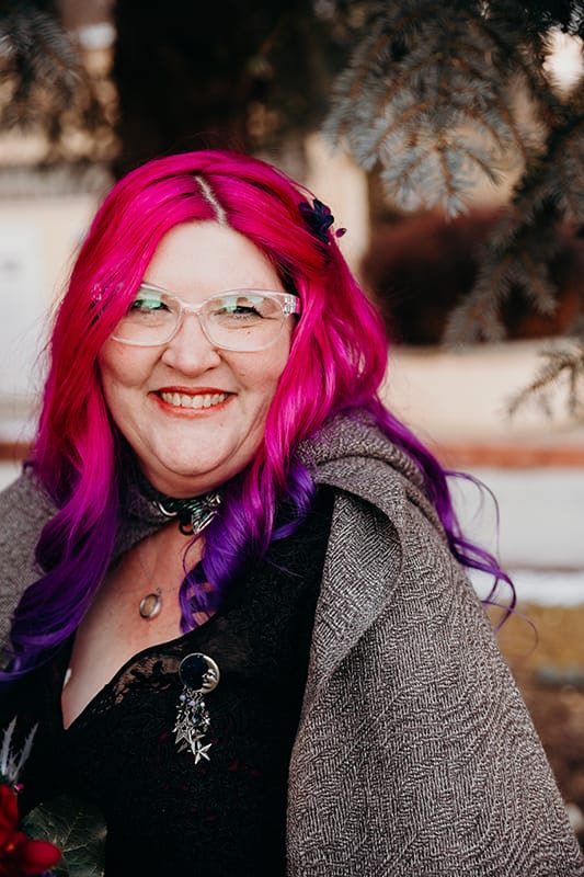 A smiling woman with glasses and pink and purple hair. Dressed in a black dress with a gray wool cloak fastened at her neck with a green leaf pin.